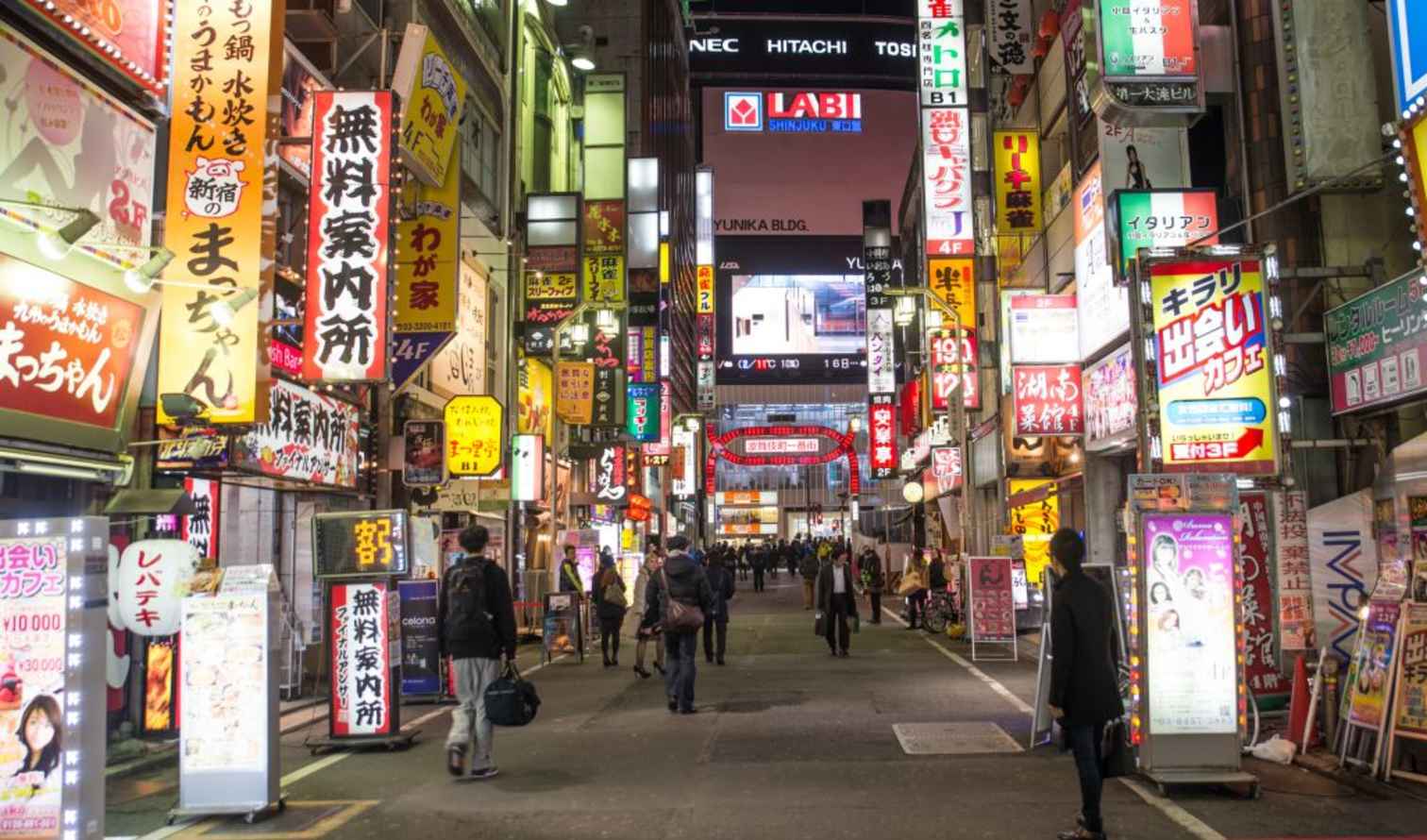 Street view of Shinjuku, Tokyo with numerous illuminated signs and people walking.