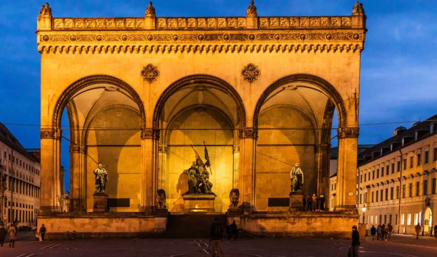 Odeonsplatz Munich lit up at dusk with statues under arches.