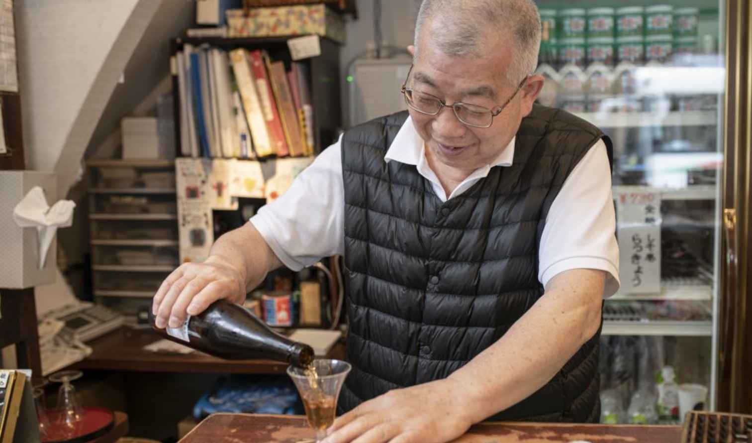 Man pours drink in a small Japanese shop with shelves behind in Tokyo
