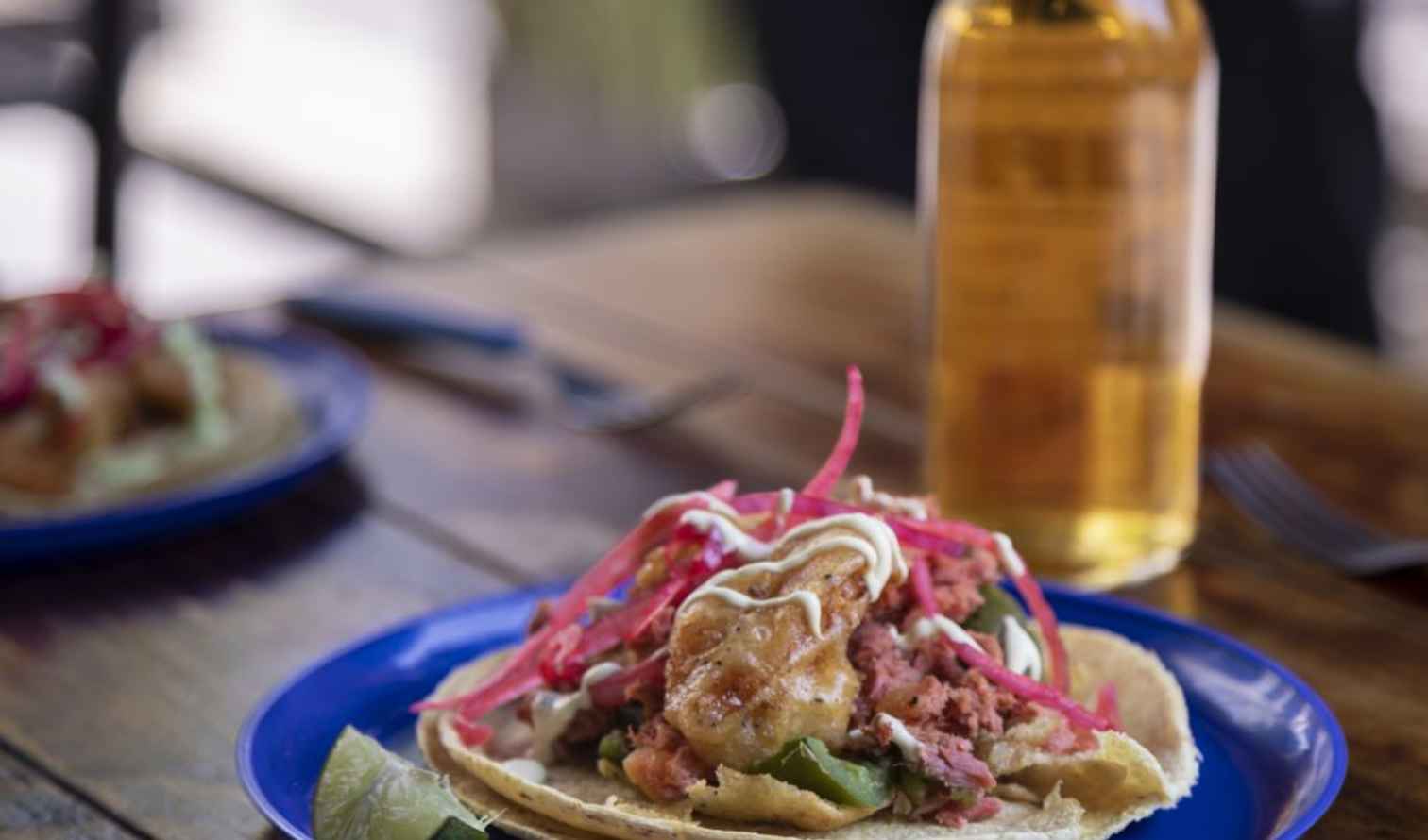 Fish taco with toppings on a blue plate beside a bottle of beer in Mexico City