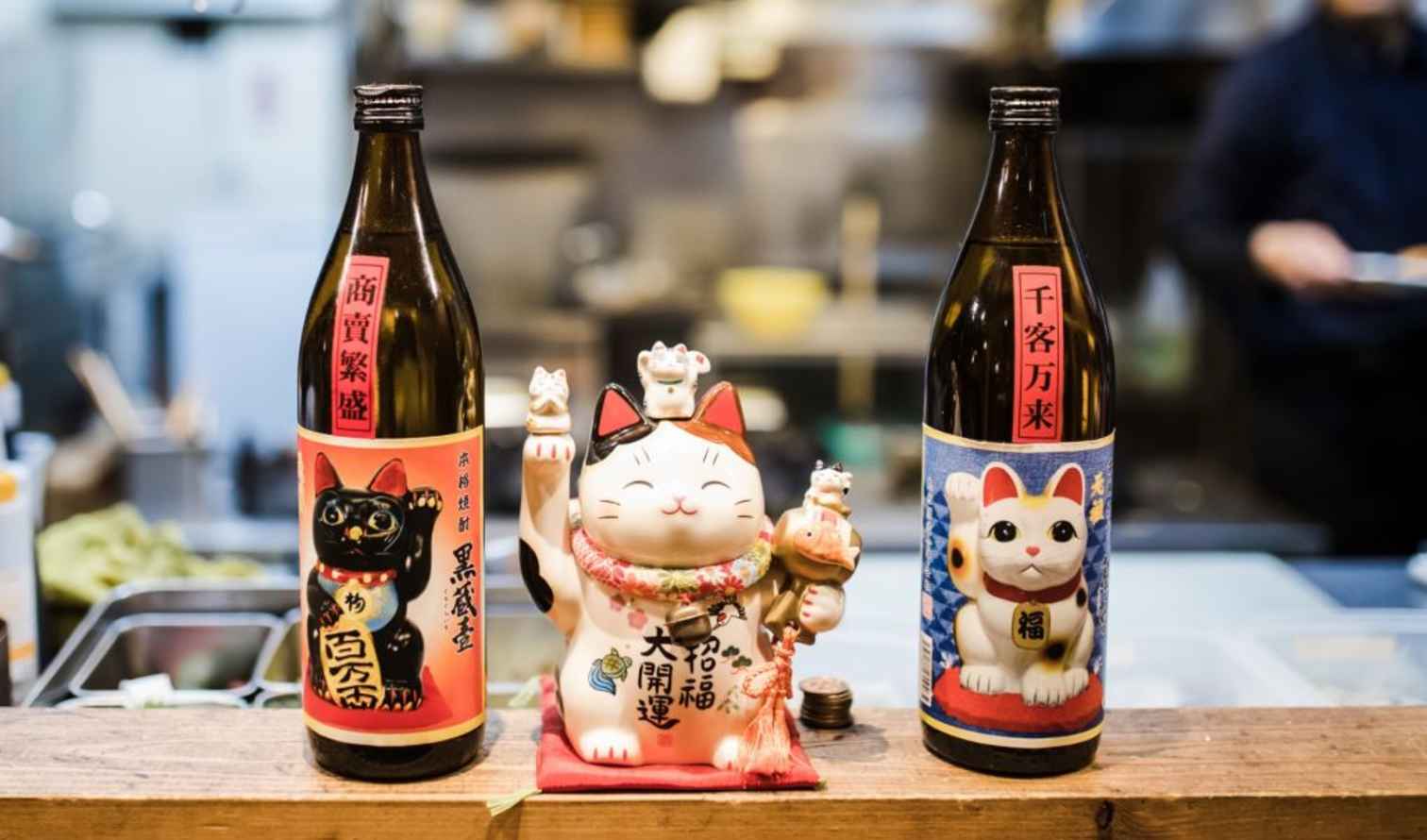 Two bottles with Maneki-neko design beside a Maneki-neko figurine on a counter in Tokyo