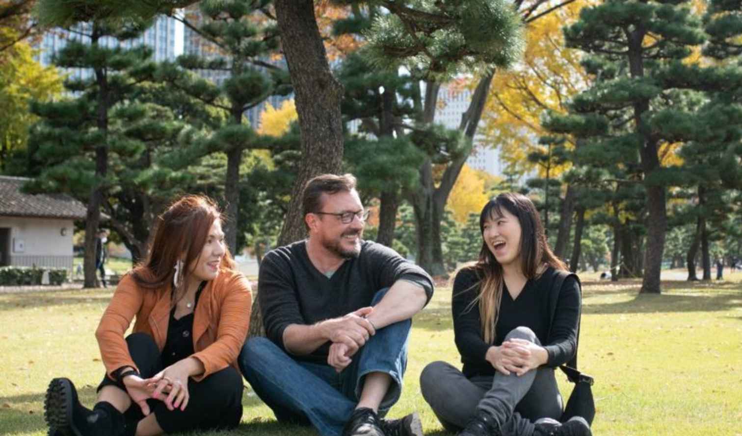 Three people sitting on grass in a park with pine trees in Tokyo