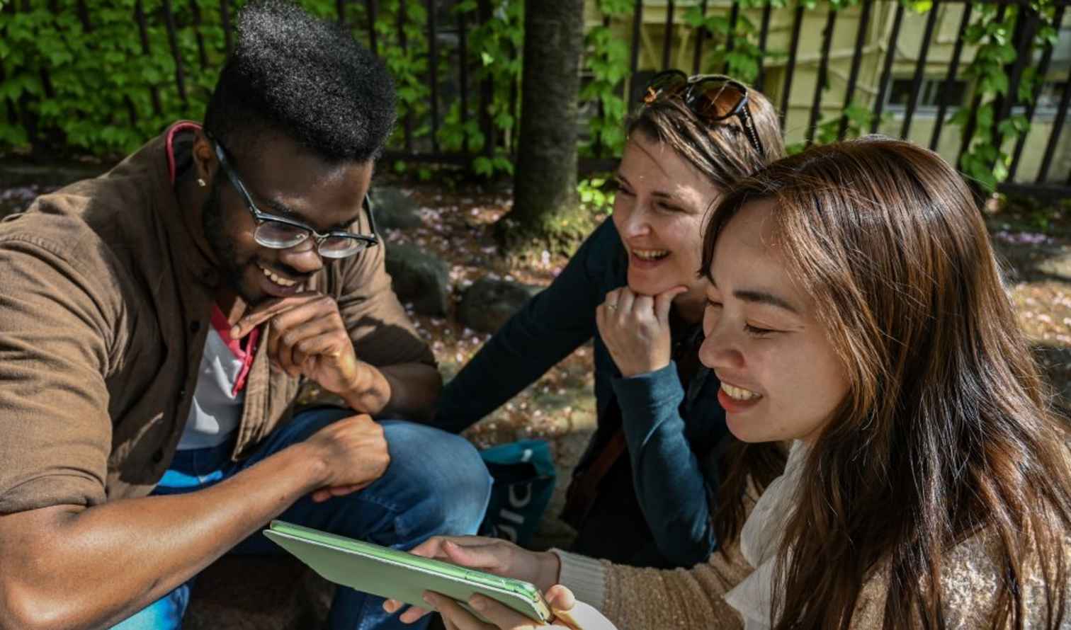 Three people looking at a tablet outdoors near a wooden fence in Kyoto