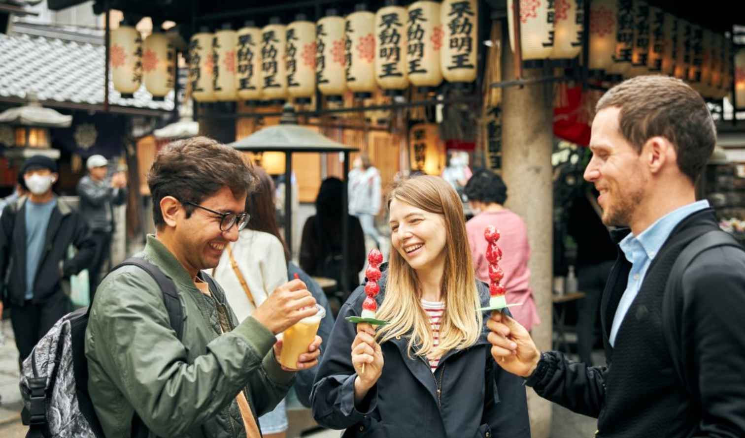 Three people holding snacks in front of Japanese lanterns in a street market in Osaka