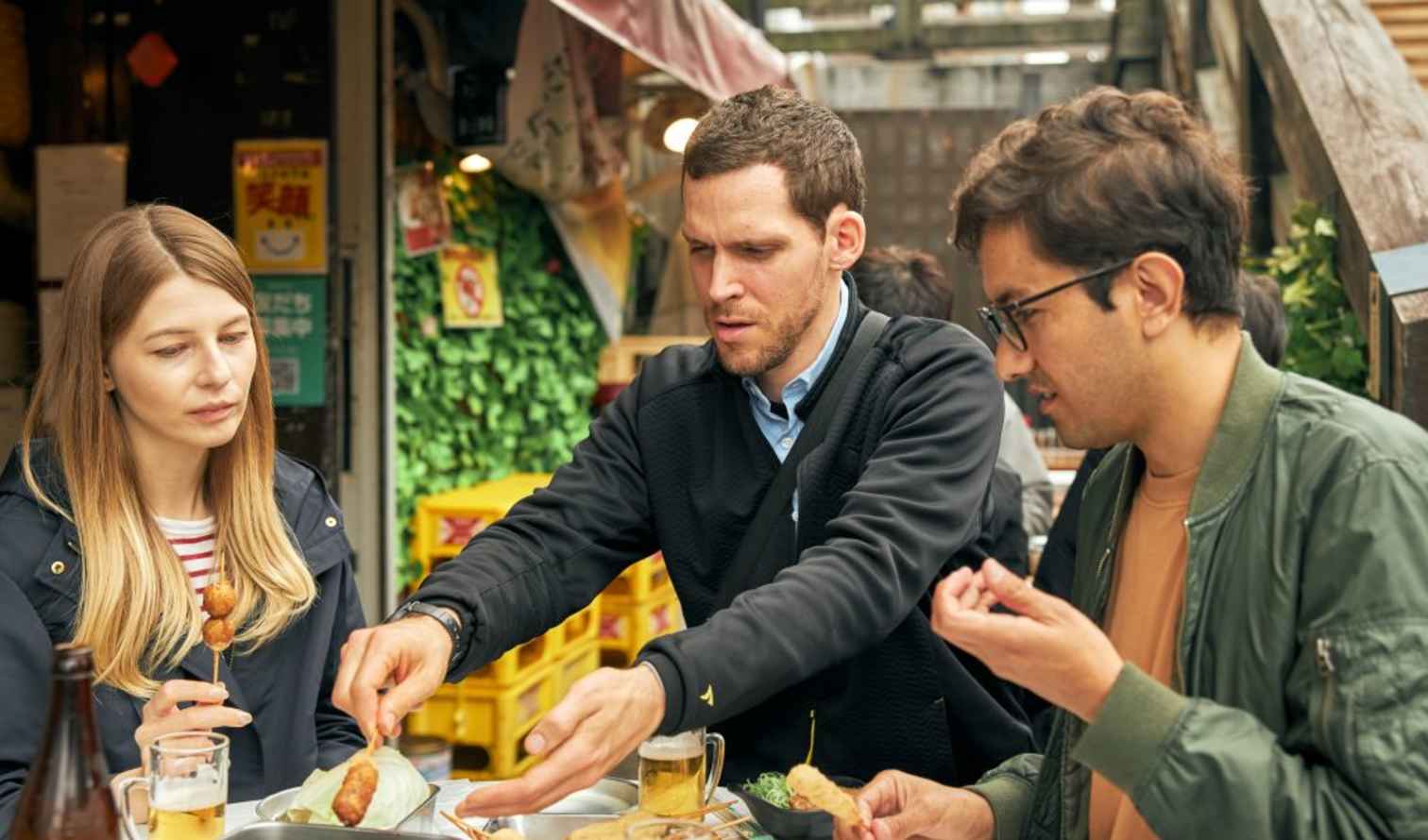 Three people sharing skewered food at a street vendor in Osaka