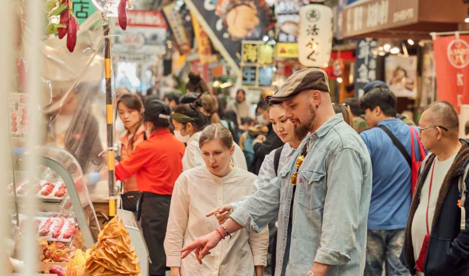 People browsing stalls at Kuromon Ichiba Market, Osaka, with street signs overhead.