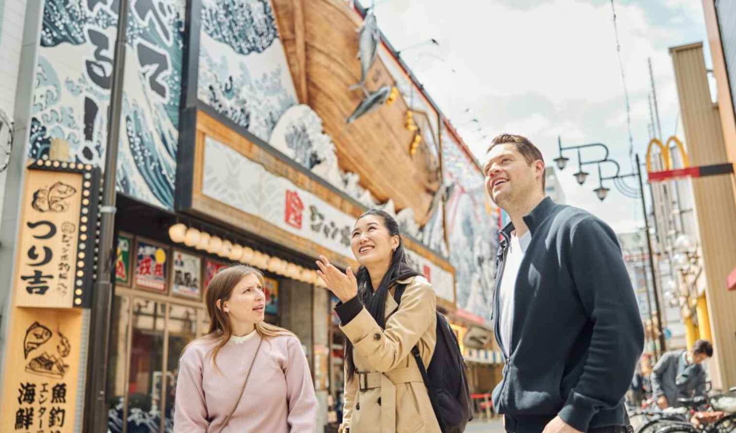 Three people walking in Dotonbori, Osaka, near a building with illustrated waves.