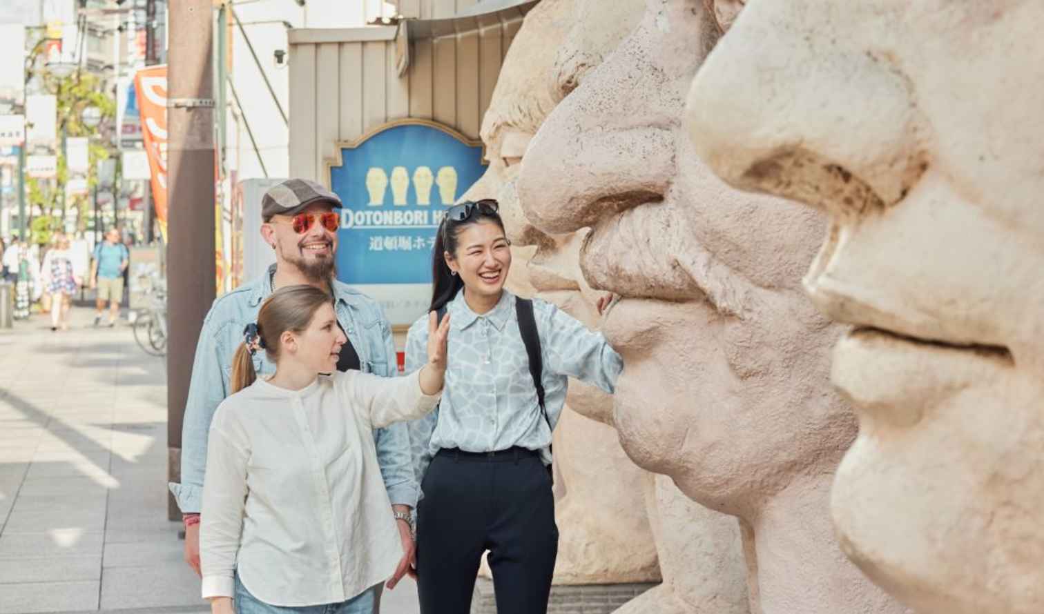 Three people stand next to large face sculptures in Dotonbori, Osaka.