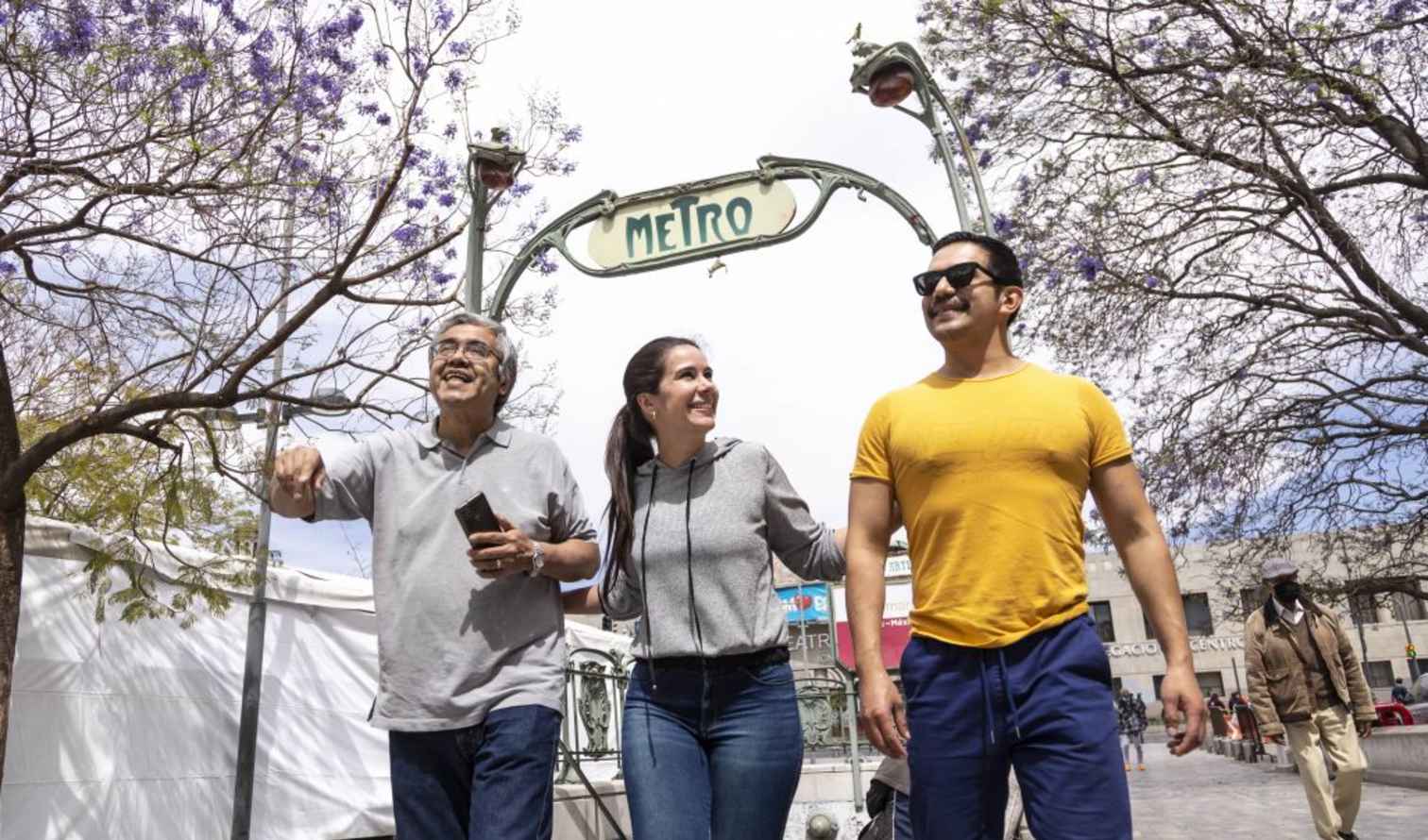 Two men and a woman near Metro sign under purple-flowered trees in  Mexico City.
