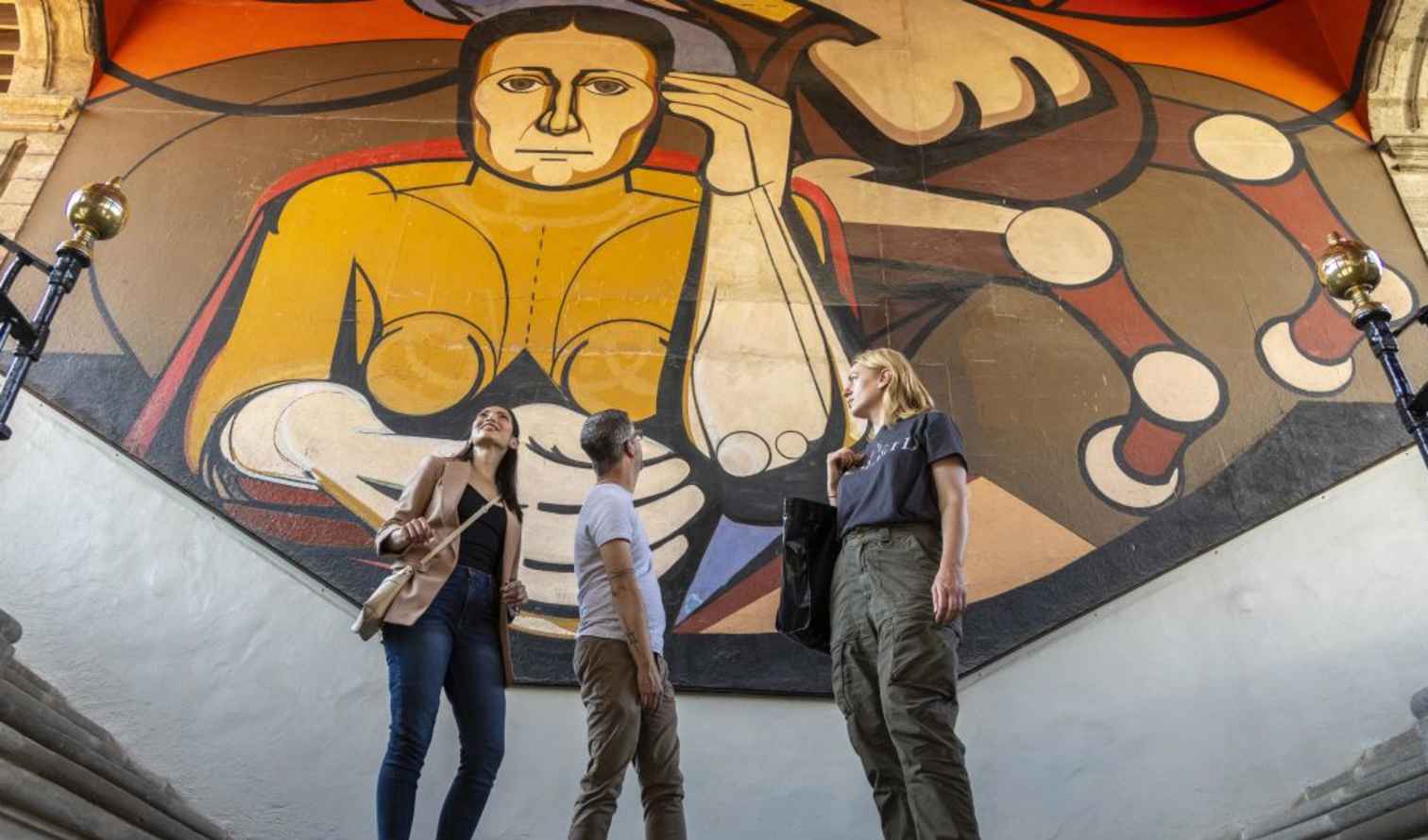 Three people standing under a large mural at Museo Casa de la Bola, Mexico City.