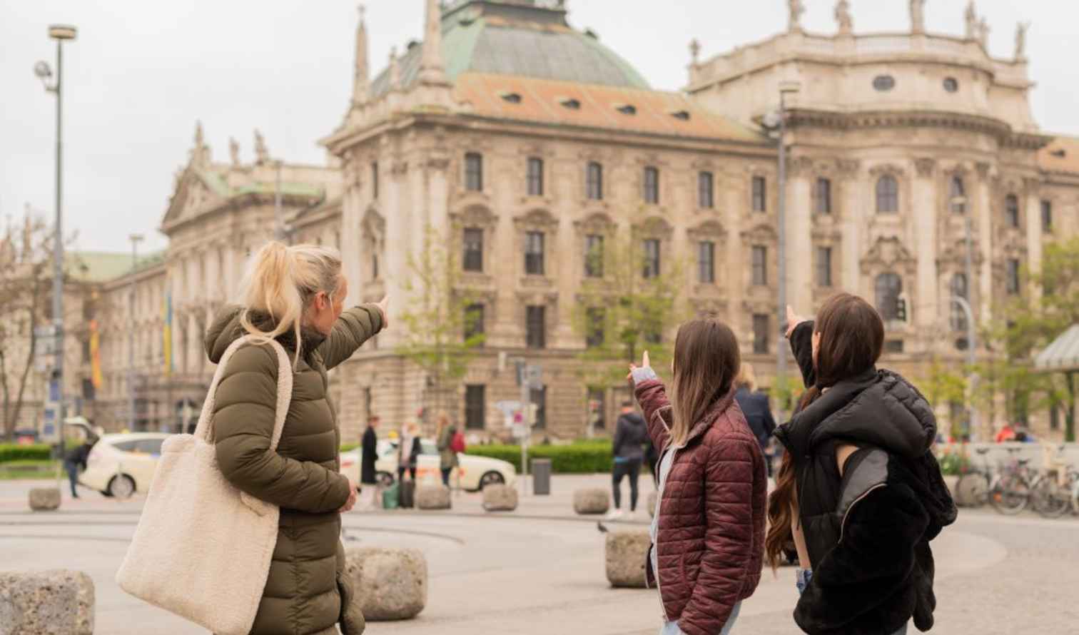 Three people pointing at the Munich courthouse building in Karlsplatz.