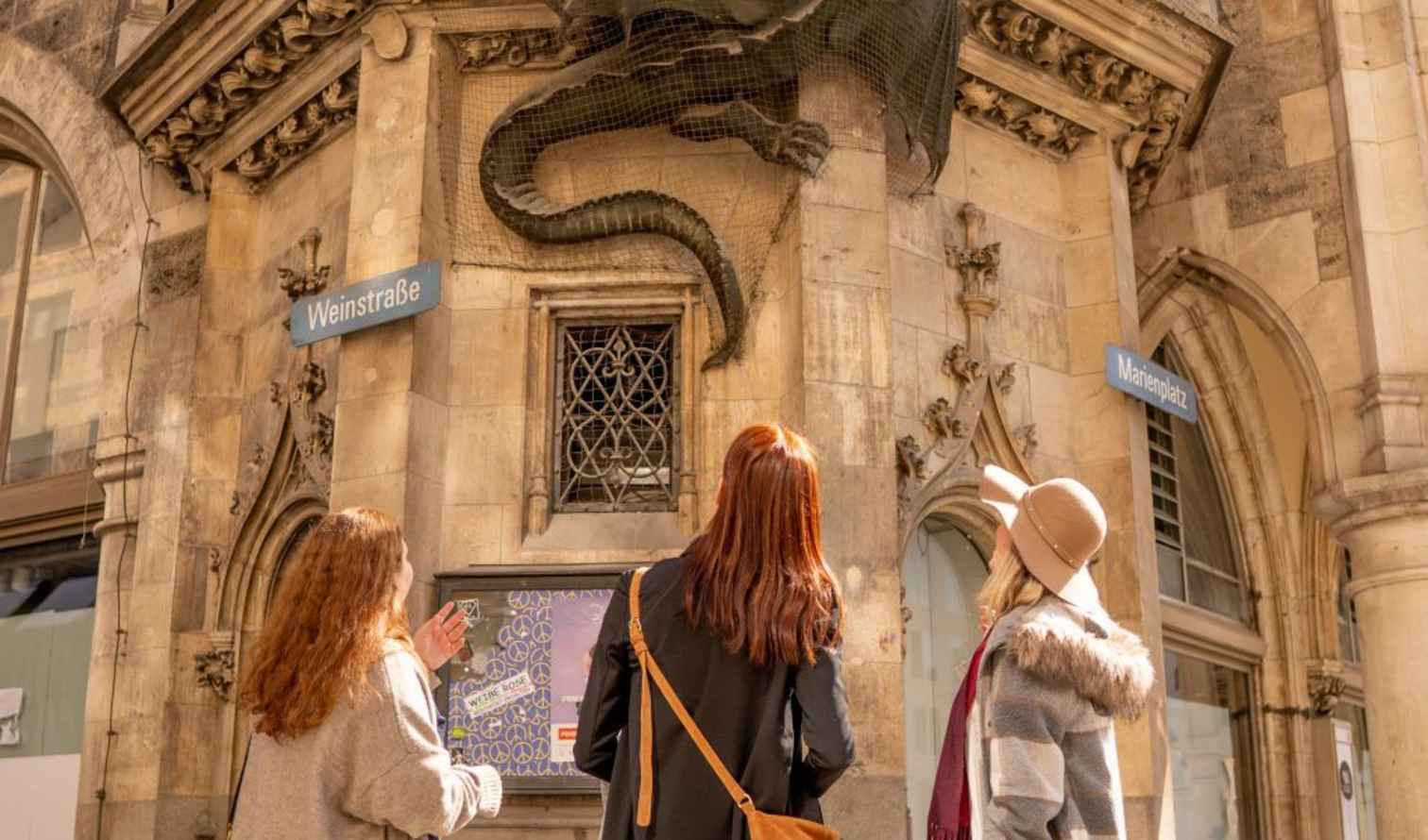Three people observing a dragon sculpture near Weinstraße sign in Munich.