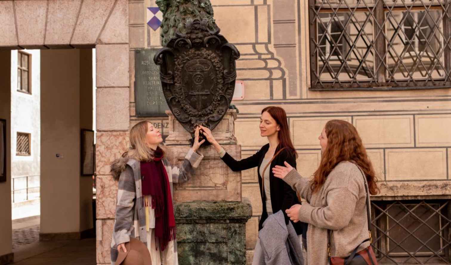 Three women touch a shield-shaped plaque in a historic European street in Munich