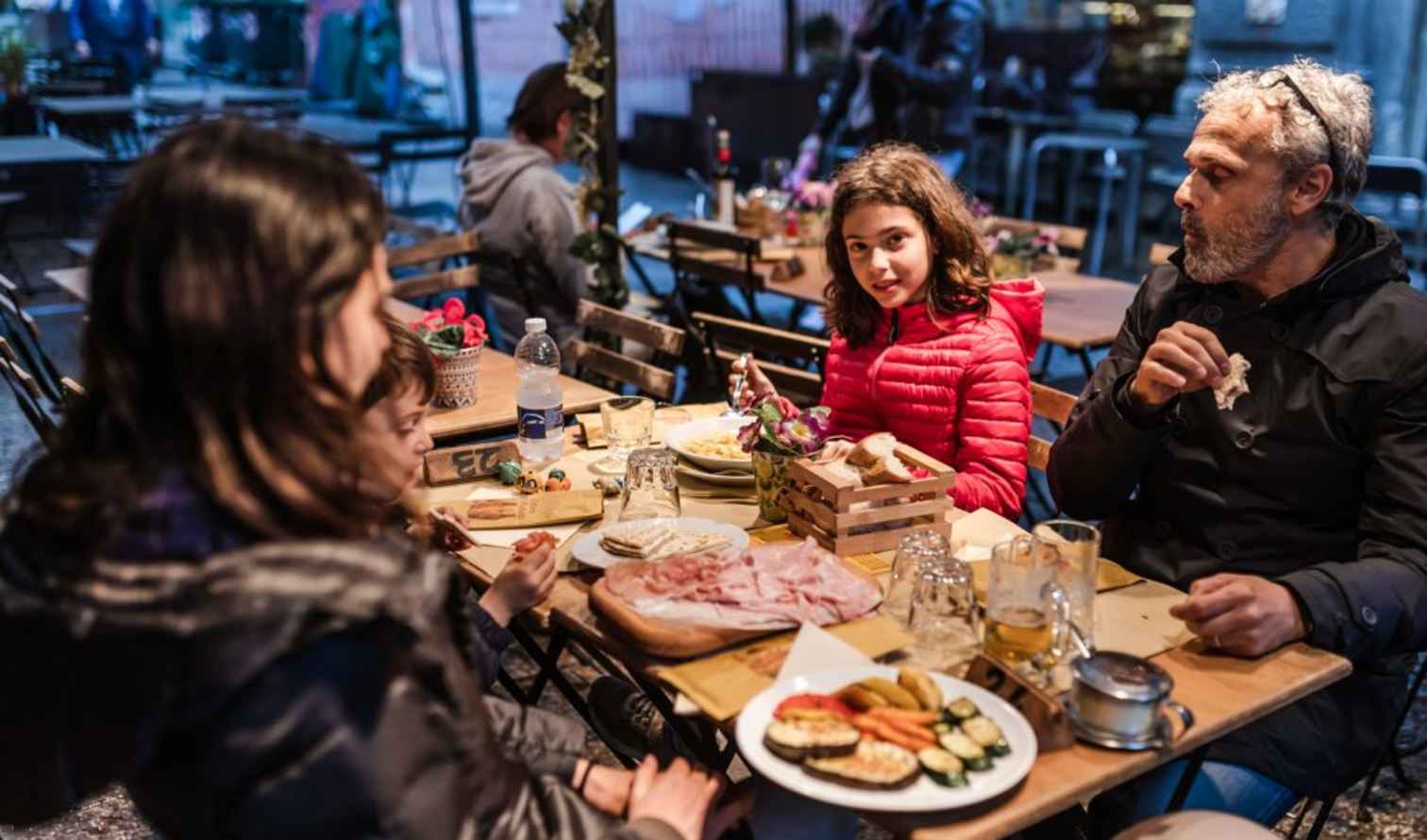 Family dining at an outdoor café with wooden tables and benches in Bologna