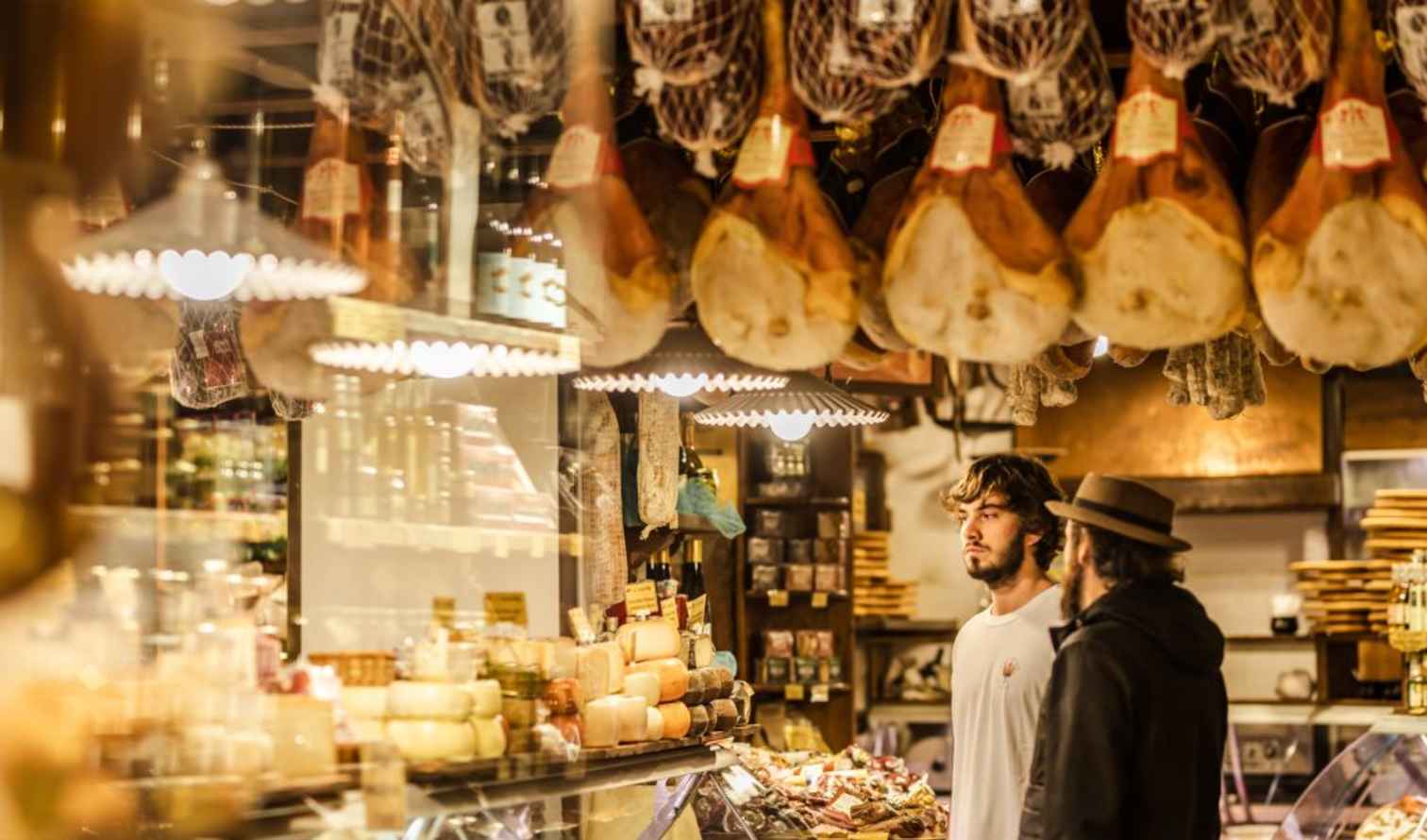 Two people browsing a deli counter with hanging cured meats in Bologna.
