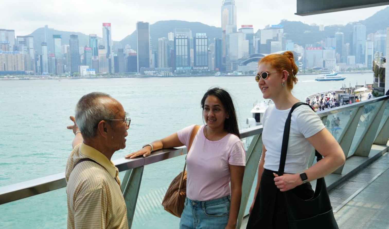Three people talking on a promenade overlooking the Hong Kong skyline.