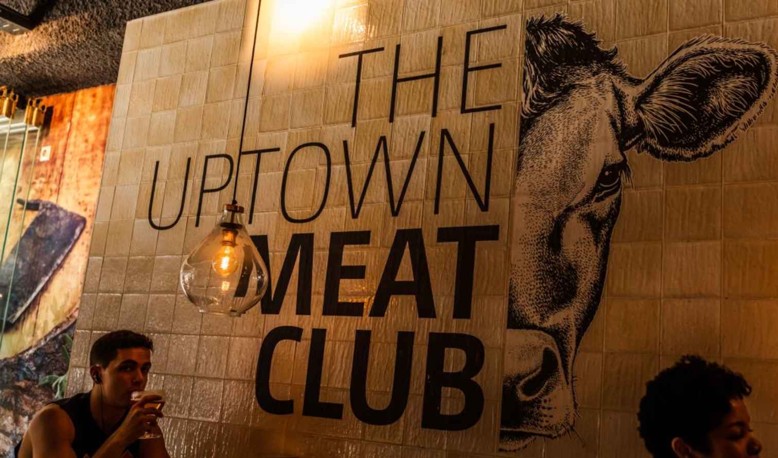 Person drinking under a light bulb beside 'The Uptown Meat Club' sign in Amsterdam.