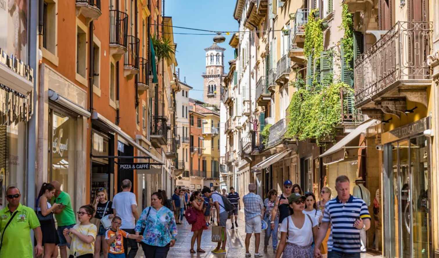 People walking on Via Mazzini in Verona, Italy, with visible tower in the background.