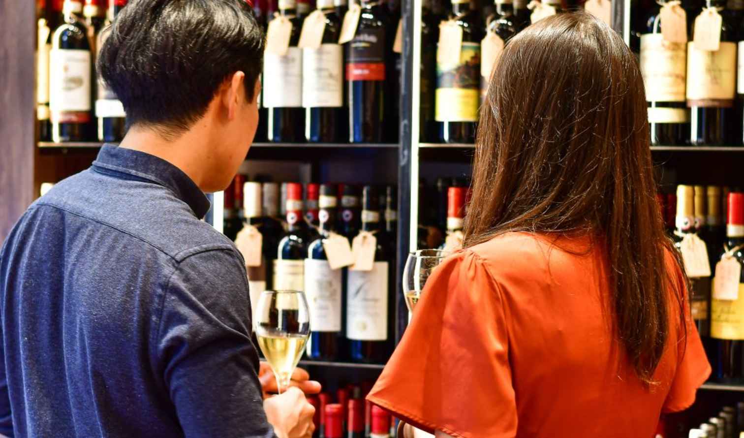 Two people hold wine glasses in front of a wine shelf in Palermo
