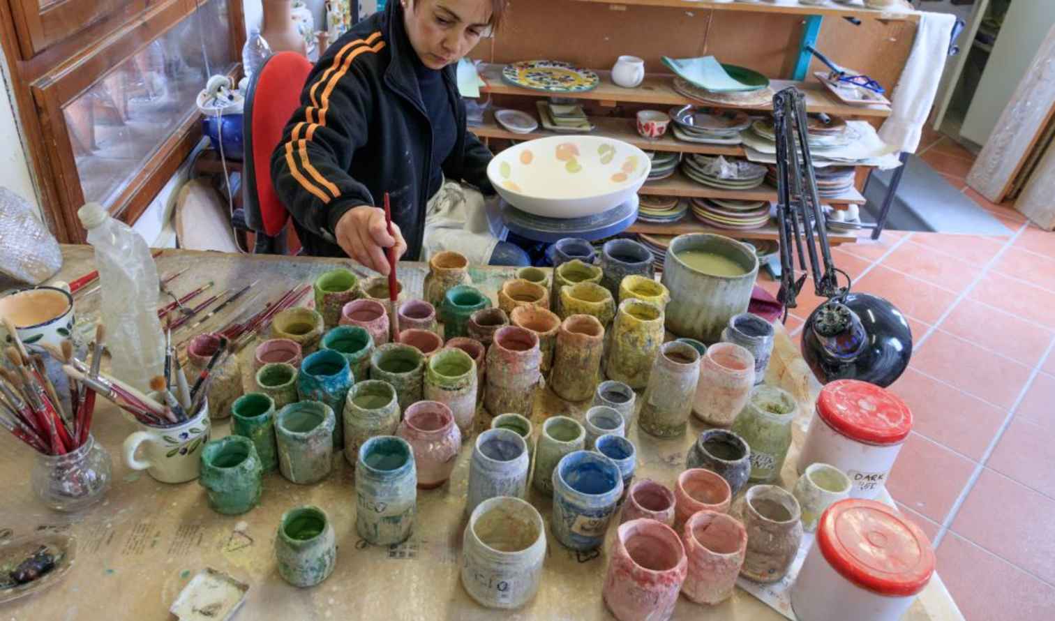 Artist painting ceramics in a workshop filled with paint jars in Sienna