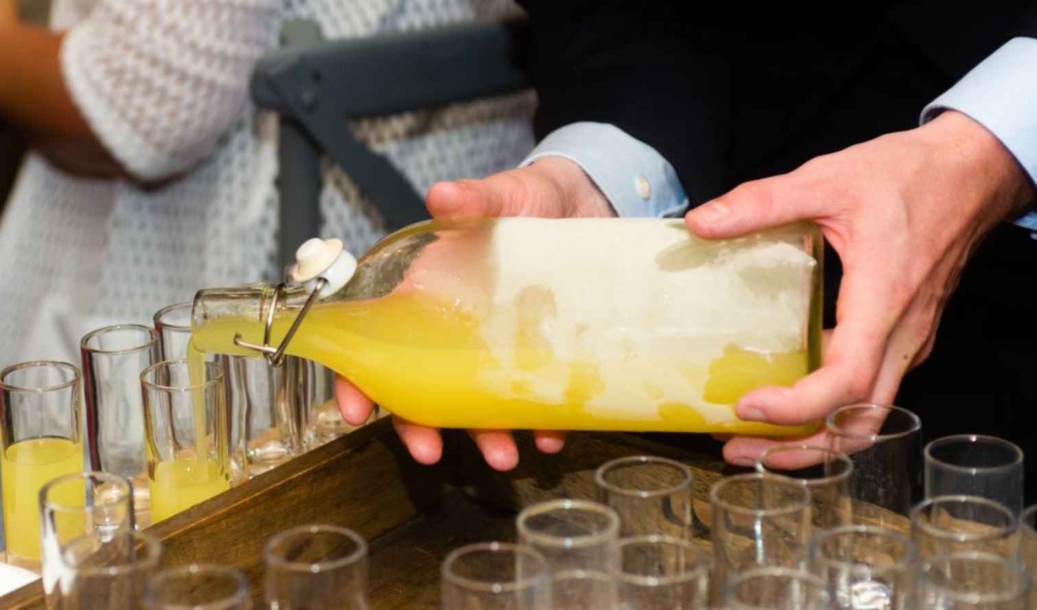 Person pouring yellow liquid from a glass bottle into several small glasses in Sorrento