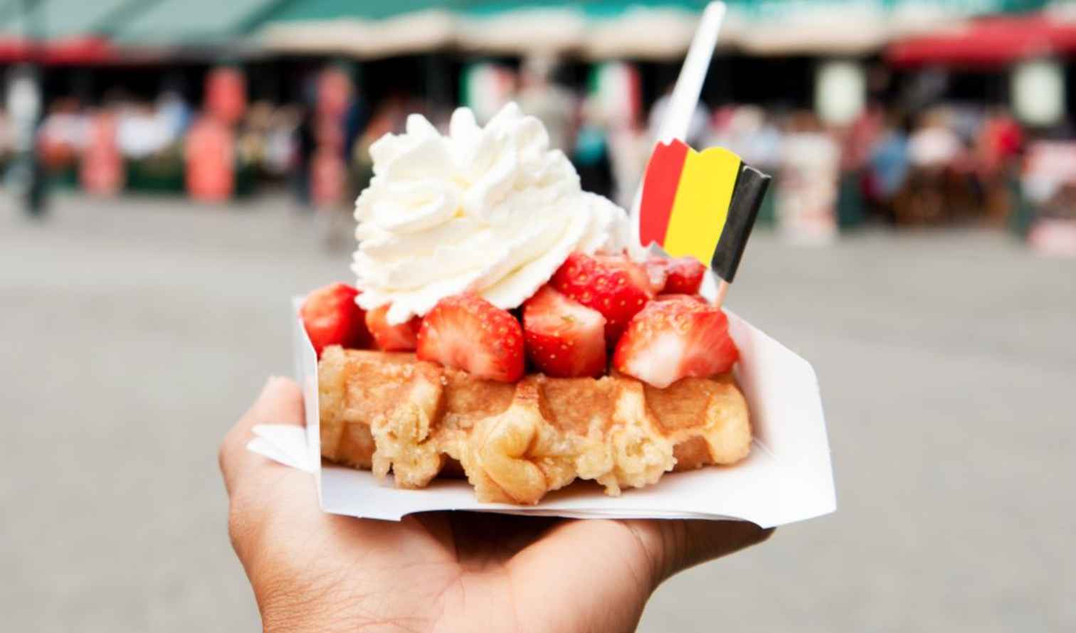 Hand holding Belgian waffle topped with strawberries and cream, market in background.