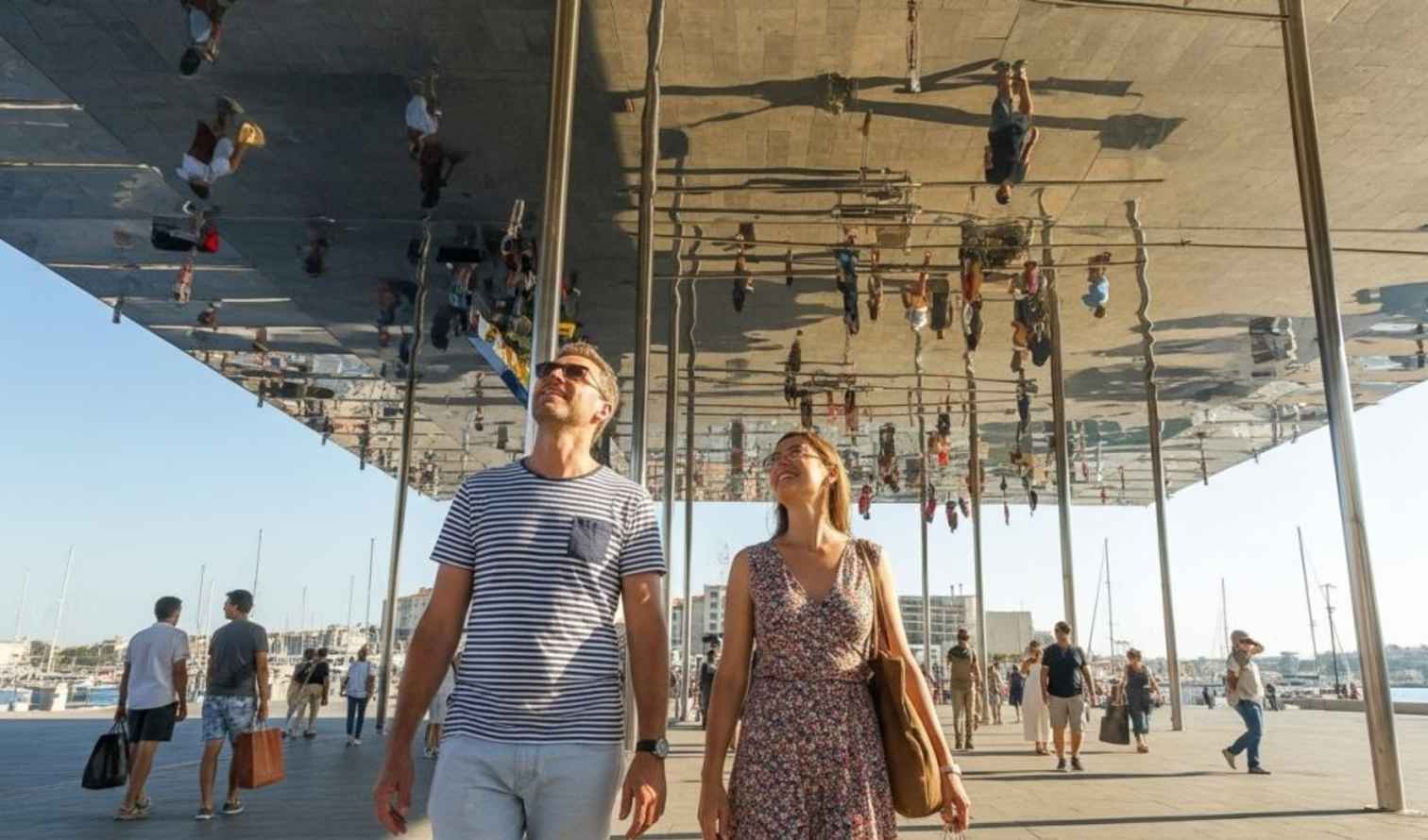 Two individuals stroll beneath the mirrored canopy at Vieux-Port, Marseille.