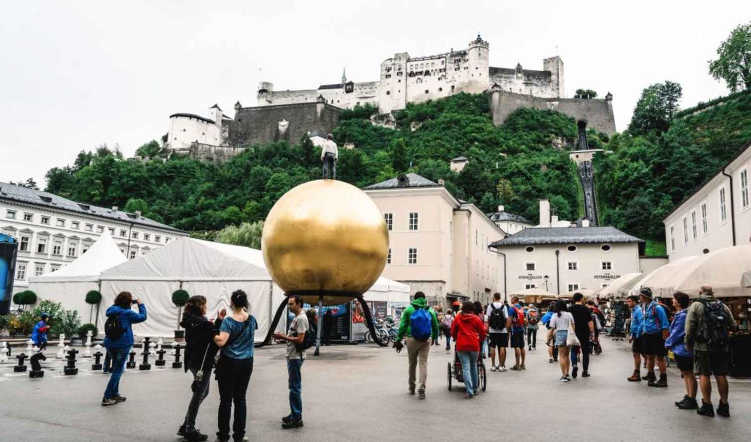 Tourists walk near a golden sculpture in Salzburg, with Hohensalzburg Fortress visible.