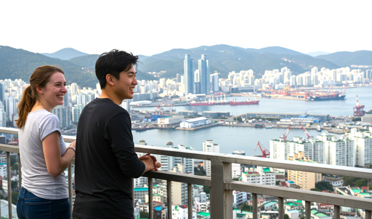 Two people view Busan's skyline from an elevated viewpoint.