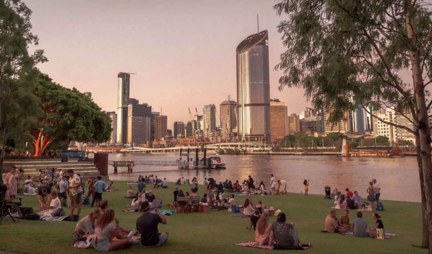People sitting on grass by the Brisbane River, skyscrapers in the background.