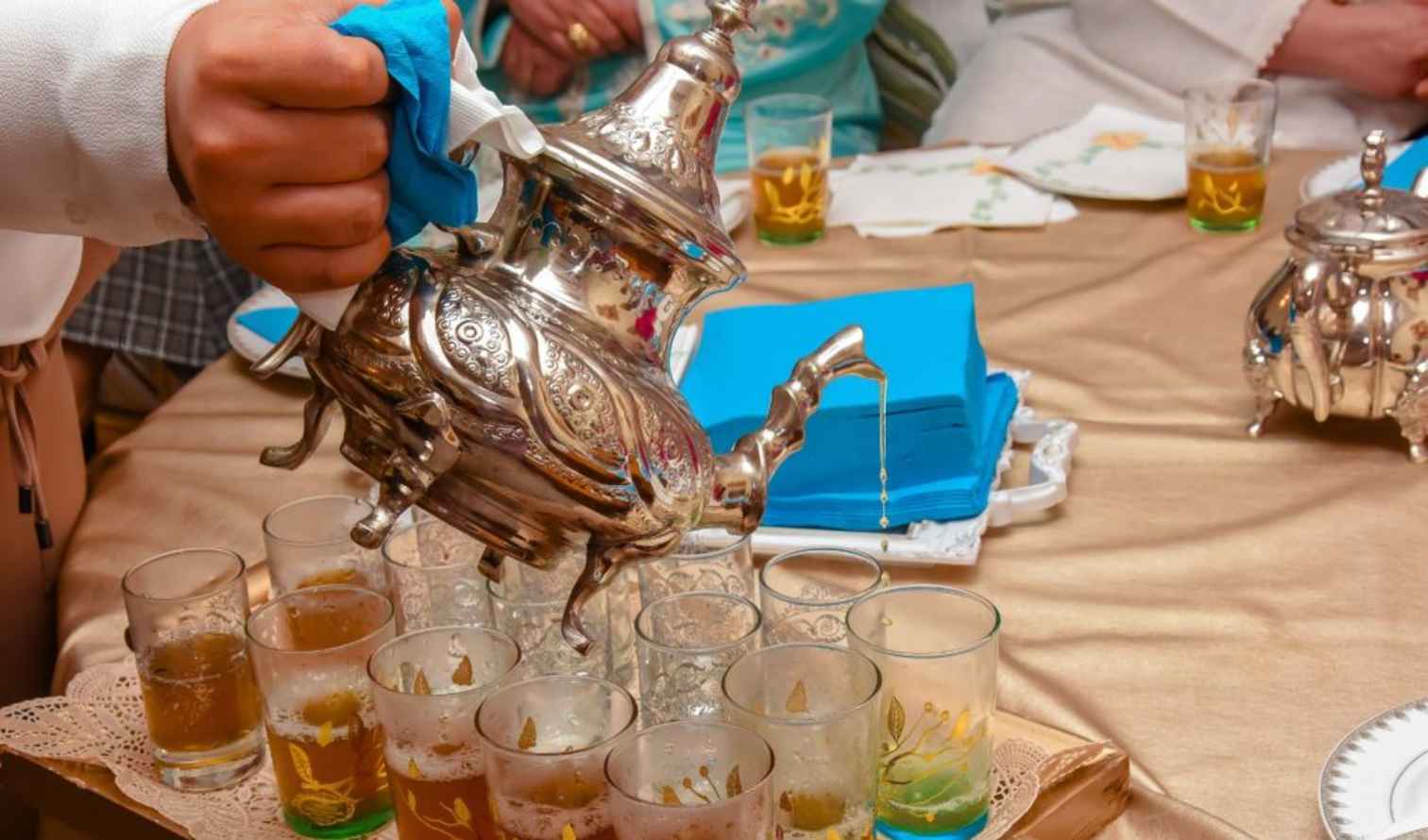 Hand pouring tea from a silver teapot into multiple glasses on a table in Marrackech.