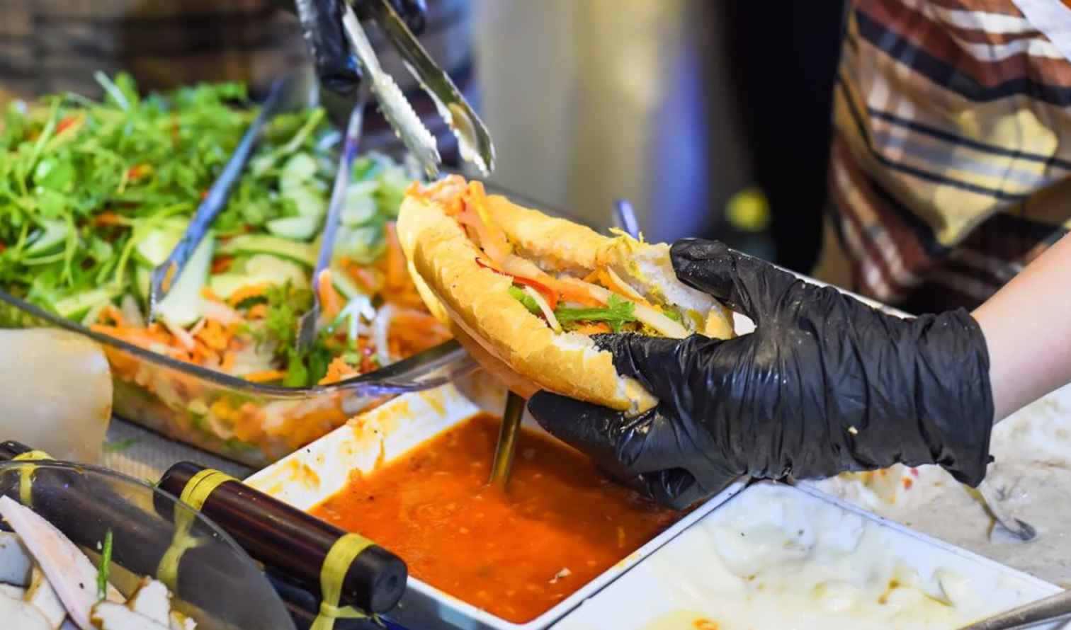 Person preparing a bánh mì sandwich with various fillings in Ho Chi Minh