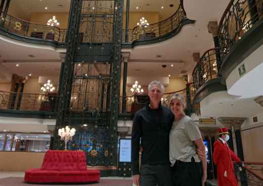 Couple standing inside a multi-story building with ornate metalwork and lighting fixtures.