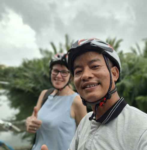 Cyclists show thumbs up next to lush greenery and a river.