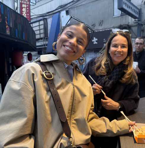 Two women with chopsticks and a food box in a Tokyo alleyway.