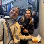 Two women with chopsticks and a food box in a Tokyo alleyway.
