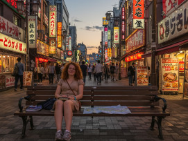 A tired but happy tourist sitting on a bench in Dotonbori at sunset, guidebook and map beside them, with the busy district continuing its evening activities in the background. A tired but happy tourist sitting on a bench in Dotonbori at sunset, guidebook and map beside them, with the busy district continuing its evening activities in the background.