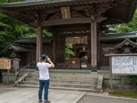 A Western visitor respectfully removing their hat while approaching a traditional Japanese temple entrance, with wooden signs displaying etiquette symbols visible nearby A Western visitor respectfully removing their hat while approaching a traditional Japanese temple entrance, with wooden signs displaying etiquette symbols visible nearby