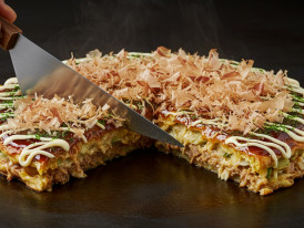 Okonomiyaki layered with mayo and bonito flakes being cut.