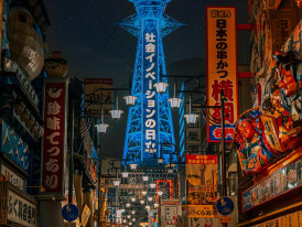 The iconic Tsutenkaku Tower glowing with neon lights against the night sky, surrounded by the bustling streets of Shinsekai district.