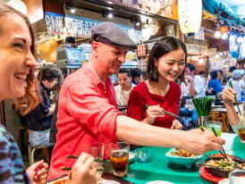 A local host and guest sharing street food from a small vendor stall.