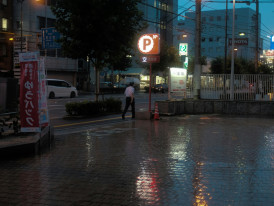 Warm sunset light reflects off rain-soaked pavement in Shinsekai, creating a moody and atmospheric street scene.  Image by Mak on Unsplash.