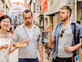 A host and guests sharing freshly made takoyaki at a lively local street food stall, laughing and enjoying the casual atmosphere.