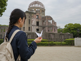 A tourist standing silently by the Atomic Bomb dome, holding a paper crane.