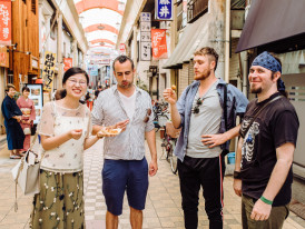 A host chatting casually with guests beside a sizzling takoyaki grill in a narrow side street.