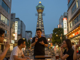 Guests listening attentively to a host sharing stories in Tsutenkaku's shadow with the iconic tower in the background.