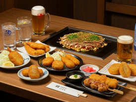 A colorful spread of kushikatsu, okonomiyaki, and local beverages on a traditional wooden table. 