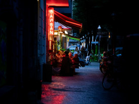 A narrow alley in Ura Namba with small bars and restaurants, their warm light spilling onto the street where a few locals chat outside. Photo by Rapha Wilde on Unsplash