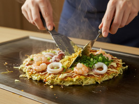 Close-up of hands mixing okonomiyaki batter with cabbage, seafood, and green onions on a table grill, showing the DIY nature of the dish