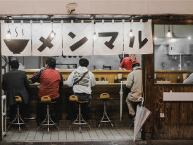 Late-night ramen stand with neon lighting and a few customers eating at the counter.